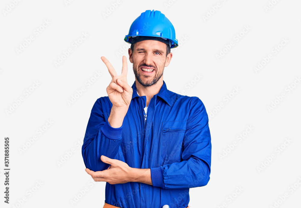 Young handsome man wearing worker uniform and hardhat smiling with happy face winking at the camera doing victory sign with fingers. number two.