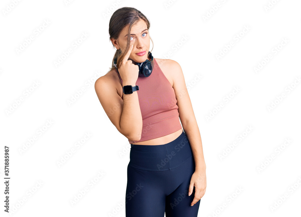 Beautiful caucasian young woman wearing gym clothes and using headphones pointing to the eye watching you gesture, suspicious expression
