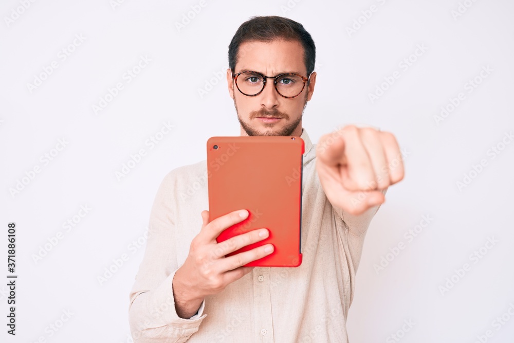 Young handsome man using touchpad device pointing with finger to the camera and to you, confident gesture looking serious