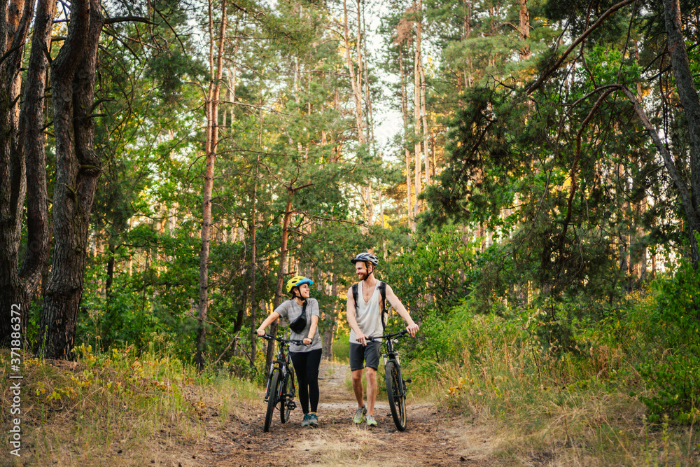 Young caucasian couple of cyclists walk and push their mountain bikes along the forest road in the park. Active sports weekend. Sporty beautiful people with a bike on the countryside