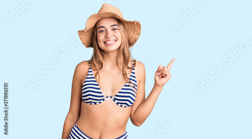 Young beautiful blonde woman wearing bikini and hat with a big smile on face, pointing with hand finger to the side looking at the camera.