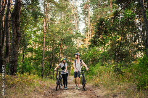 Young caucasian couple of cyclists walk and push their mountain bikes along the forest road in the park. Active sports weekend. Sporty beautiful people with a bike on the countryside