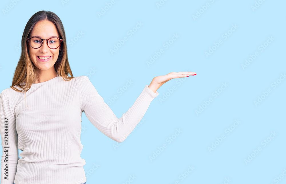 Beautiful brunette young woman wearing casual white sweater and glasses smiling cheerful presenting and pointing with palm of hand looking at the camera. Beautiful brunette young woman wearing casual white sweater and glasses smiling cheerful presenting and pointing with palm of hand looking at the camera.