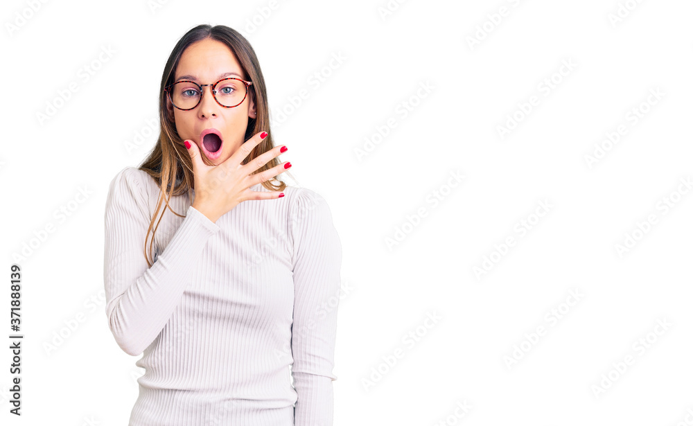 Beautiful brunette young woman wearing casual white sweater and glasses looking fascinated with disbelief, surprise and amazed expression with hands on chin Beautiful brunette young woman wearing casual white sweater and glasses looking fascinated with disbelief, surprise and amazed expression with hands on chin