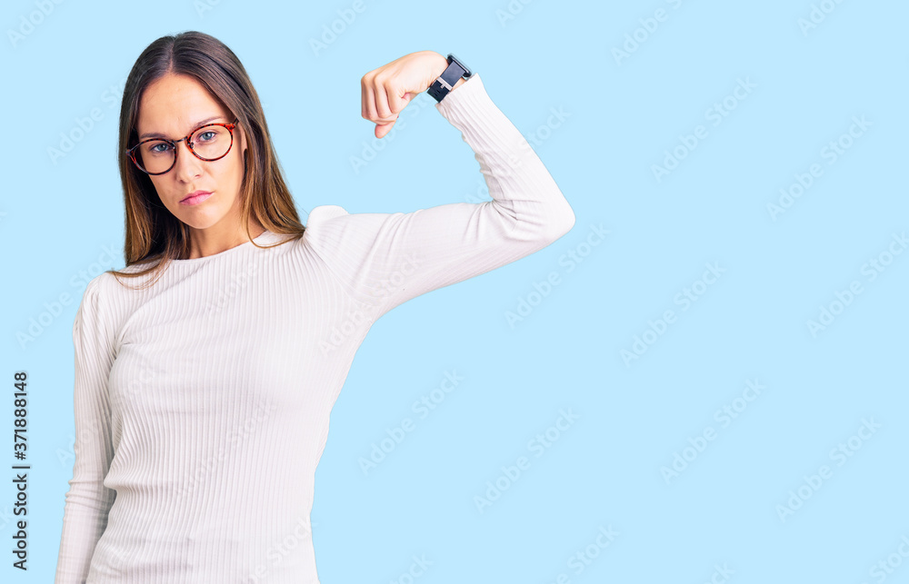 Beautiful brunette young woman wearing casual white sweater and glasses strong person showing arm muscle, confident and proud of power Beautiful brunette young woman wearing casual white sweater and glasses strong person showing arm muscle, confident and proud of power