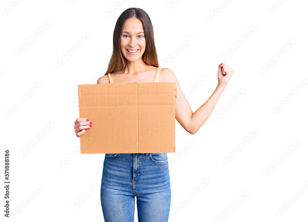 Beautiful brunette young woman holding cardboard banner with blank space screaming proud, celebrating victory and success very excited with raised arms Beautiful brunette young woman holding cardboard banner with blank space screaming proud, celebrating victory and success very excited with raised arms