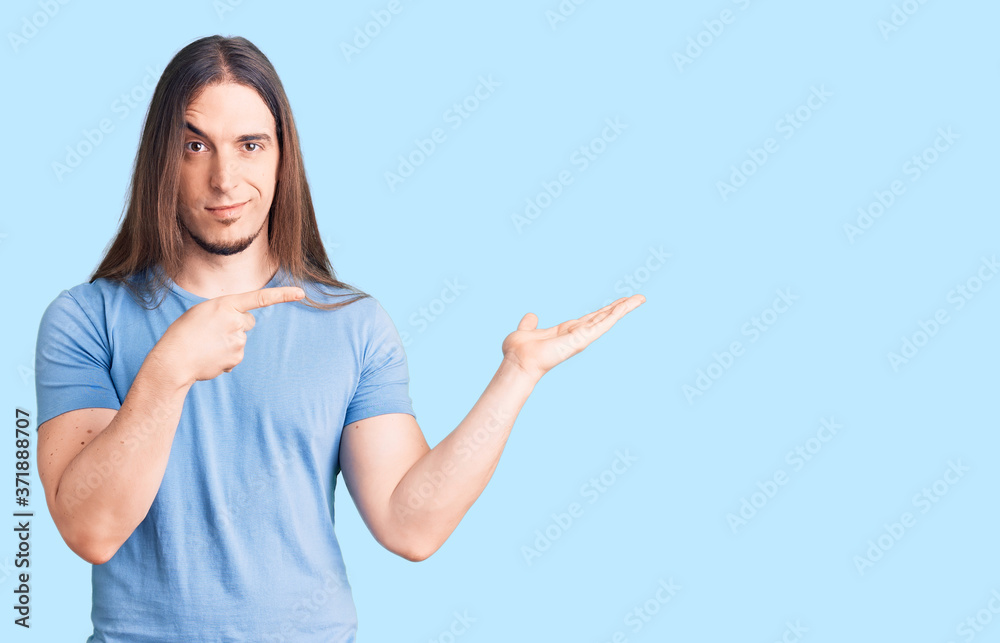 Young adult man with long hair wearing casual clothes amazed and smiling to the camera while presenting with hand and pointing with finger. Young adult man with long hair wearing casual clothes amazed and smiling to the camera while presenting with hand and pointing with finger.