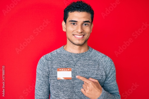 Young handsome hispanic man wearing hello my name is sticker identification smiling happy pointing with hand and finger photo