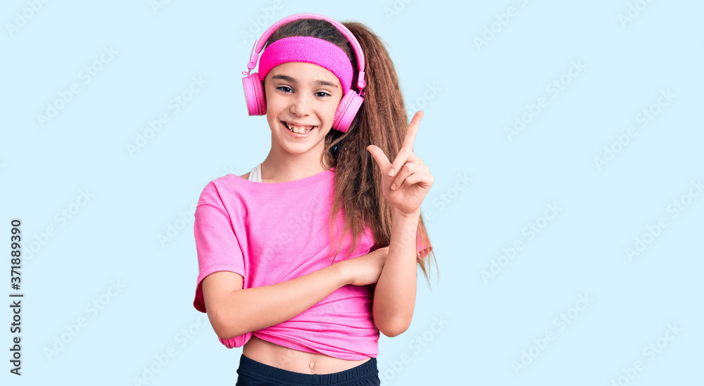 Cute hispanic child girl wearing gym clothes and using headphones with a big smile on face, pointing with hand and finger to the side looking at the camera. Cute hispanic child girl wearing gym clothes and using headphones with a big smile on face, pointing with hand and finger to the side looking at the camera.