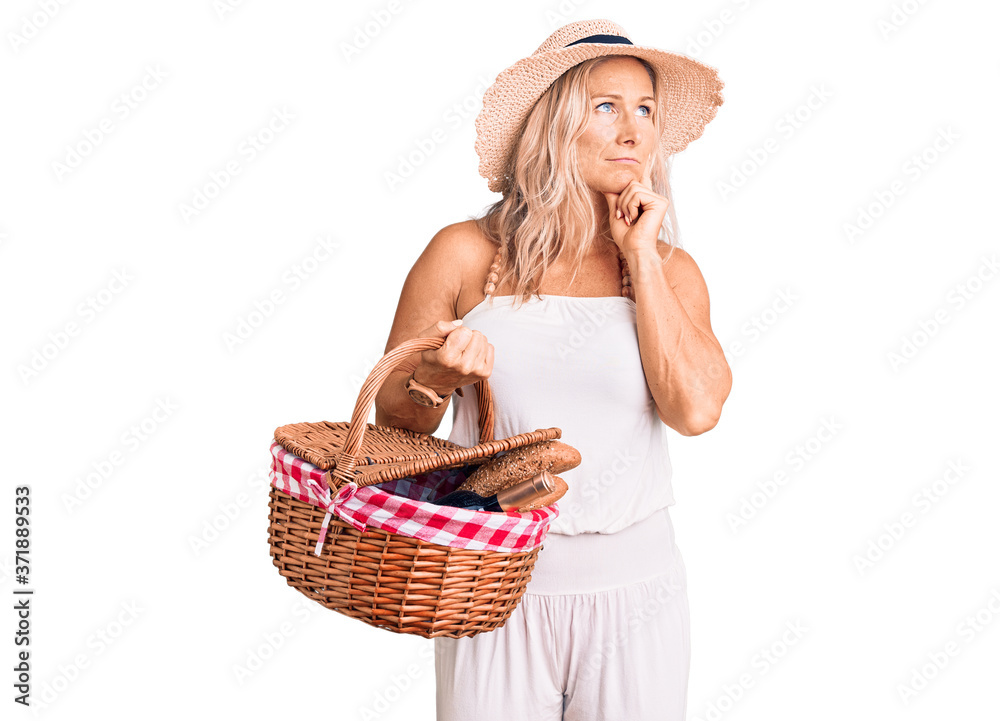 Middle age fit blonde woman wearing summer hat and holding picnic wicker basket with bread serious face thinking about question with hand on chin, thoughtful about confusing idea Middle age fit blonde woman wearing summer hat and holding picnic wicker basket with bread serious face thinking about question with hand on chin, thoughtful about confusing idea