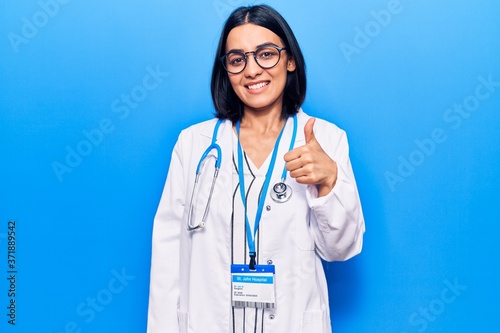 Young beautiful latin woman wearing doctor stethoscope and id card smiling happy and positive, thumb up doing excellent and approval sign photo