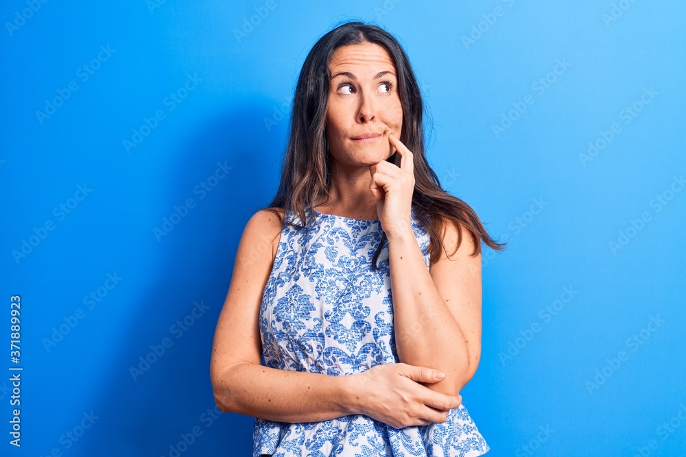 Fototapeta premium Young beautiful brunette woman wearing casual sleeveless t-shirt over blue background thinking concentrated about doubt with finger on chin and looking up wondering