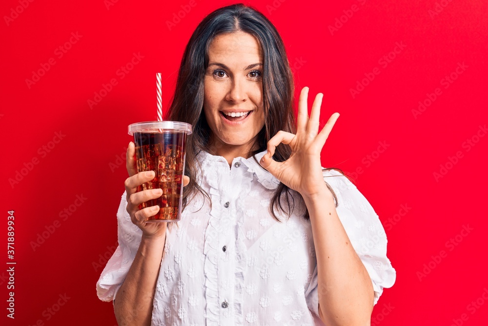 Beautiful brunette woman drinking cola refreshment beverage using straw over red background doing ok sign with fingers, smiling friendly gesturing excellent symbol