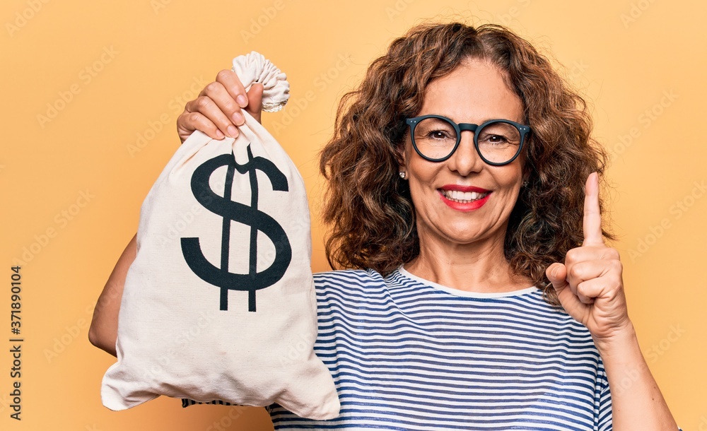 Middle age beautiful woman holding bag with dollar symbol over isolated yellow background smiling with an idea or question pointing finger with happy face, number one Middle age beautiful woman holding bag with dollar symbol over isolated yellow background smiling with an idea or question pointing finger with happy face, number one