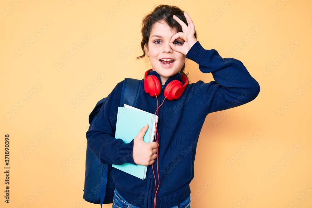 Cute hispanic child holding student backpack and books smiling happy doing ok sign with hand on eye looking through fingers Cute hispanic child holding student backpack and books smiling happy doing ok sign with hand on eye looking through fingers