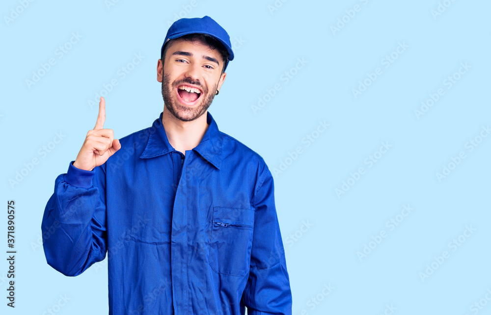 Young hispanic man wearing painter uniform pointing finger up with successful idea. exited and happy. number one.