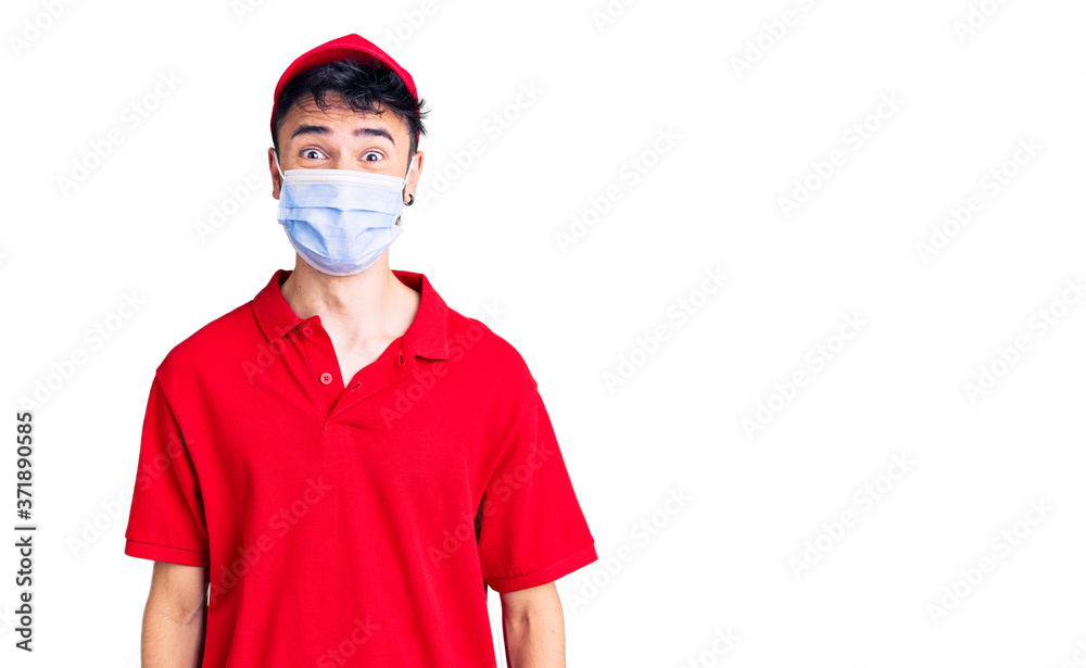 Young hispanic man wearing delivery uniform and medical mask scared and amazed with open mouth for surprise, disbelief face