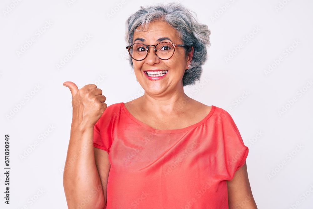 Senior hispanic grey- haired woman wearing casual clothes and glasses smiling with happy face looking and pointing to the side with thumb up. Senior hispanic grey- haired woman wearing casual clothes and glasses smiling with happy face looking and pointing to the side with thumb up.