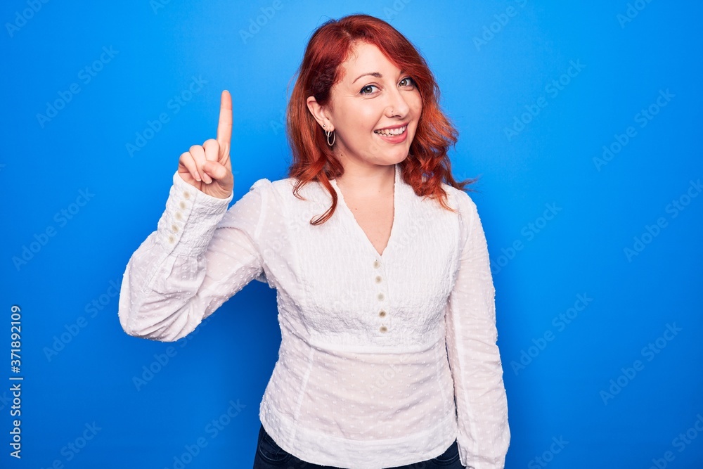 Fototapeta premium Young beautiful redhead businesswoman wearing elegant shirt standing over blue background smiling with an idea or question pointing finger up with happy face, number one