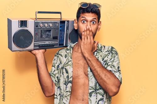 Young latin man wearing summer shirt holding boombox covering mouth with hand, shocked and afraid for mistake. surprised expression
