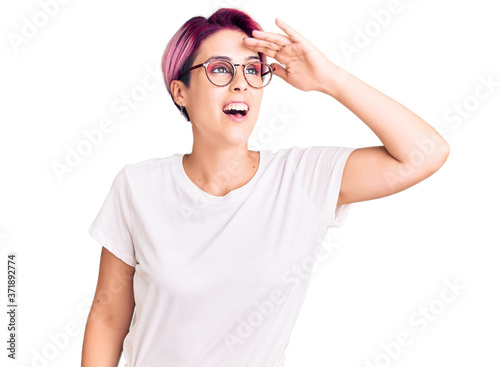 Young beautiful woman with pink hair wearing casual clothes and glasses very happy and smiling looking far away with hand over head. searching concept.