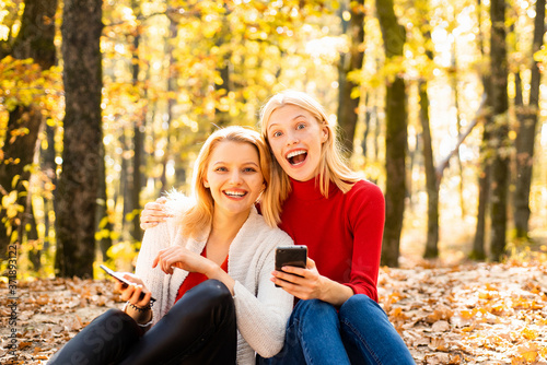 Happy young girls holding mobile phone and sitting on fallen leaves outdoor. Young women in Autumn. Students friends on beautiful fall time in nature.