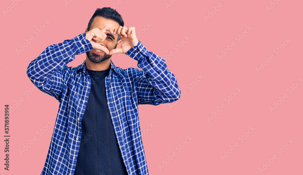 Young hispanic man wearing casual clothes doing heart shape with hand and fingers smiling looking through sign