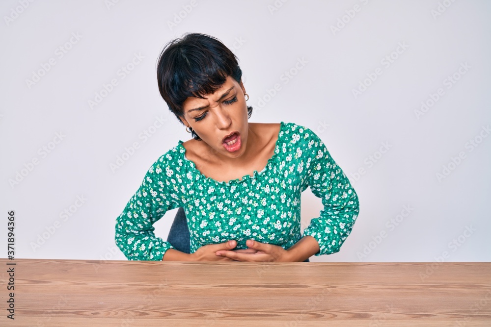 Beautiful brunettte woman wearing casual clothes sitting on the table with hand on stomach because nausea, painful disease feeling unwell. ache concept. Beautiful brunettte woman wearing casual clothes sitting on the table with hand on stomach because nausea, painful disease feeling unwell. ache concept.
