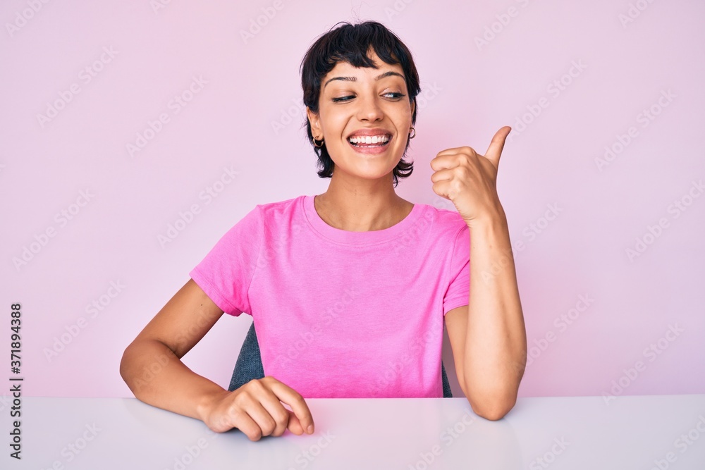Beautiful brunettte woman wearing casual clothes over pink background smiling with happy face looking and pointing to the side with thumb up. Beautiful brunettte woman wearing casual clothes over pink background smiling with happy face looking and pointing to the side with thumb up.