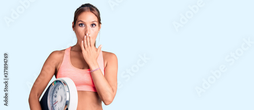 Young beautiful hispanic woman wearing sportswear holding weighing machine covering mouth with hand  shocked and afraid for mistake. surprised expression