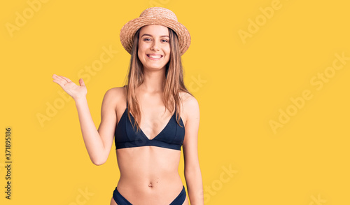 Young beautiful girl wearing bikini and hat smiling cheerful presenting and pointing with palm of hand looking at the camera.