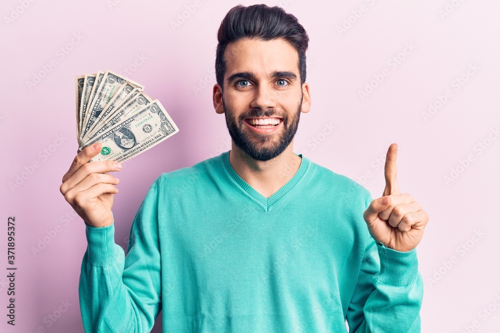 Young handsome man with beard holding bunch of dollar smiling with an idea or question pointing finger with happy face, number one Young handsome man with beard holding bunch of dollar smiling with an idea or question pointing finger with happy face, number one
