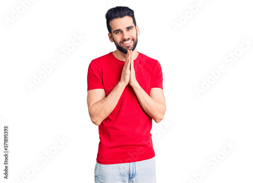 Young handsome man with beard wearing casual t-shirt praying with hands together asking for forgiveness smiling confident.