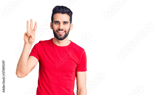 Young handsome man with beard wearing casual t-shirt showing and pointing up with fingers number three while smiling confident and happy.
