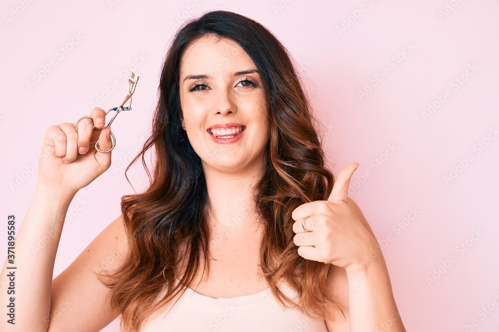 Young beautiful brunette woman holding eyelash curler smiling happy and positive, thumb up doing excellent and approval sign Young beautiful brunette woman holding eyelash curler smiling happy and positive, thumb up doing excellent and approval sign