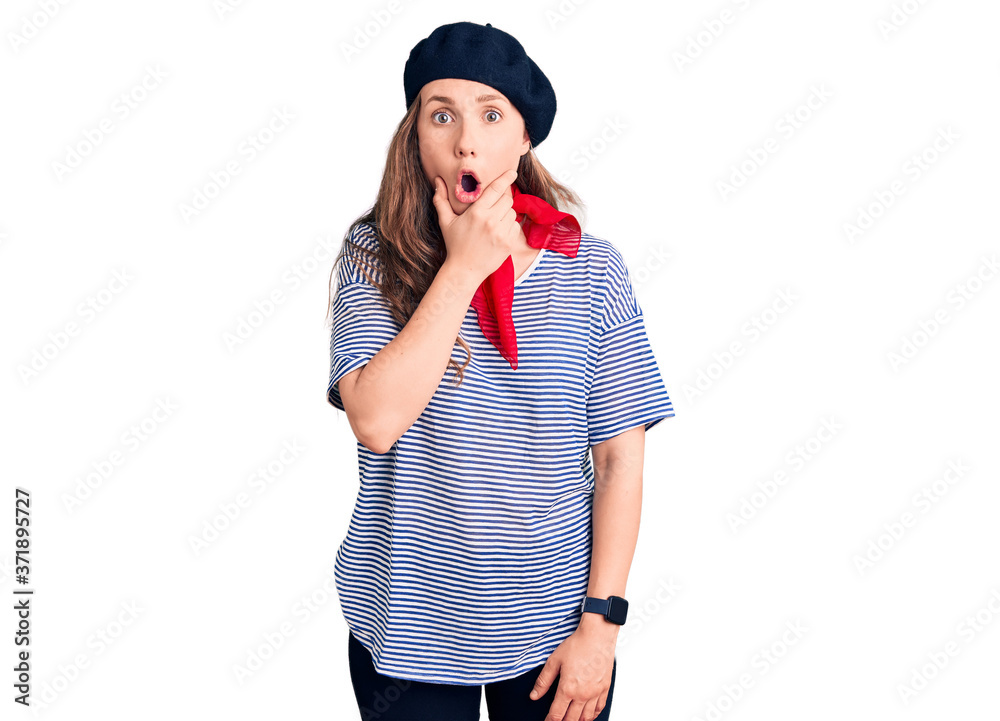 Young beautiful blonde woman wearing french beret and striped t-shirt looking fascinated with disbelief, surprise and amazed expression with hands on chin Young beautiful blonde woman wearing french beret and striped t-shirt looking fascinated with disbelief, surprise and amazed expression with hands on chin