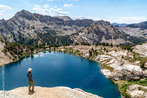 Man, hiker, standing, watching a scene of an alpine lake and rugged mountains with trees, rocks, and puffy clouds in the blue sky, Liberty Lake, Ruby Mountain Range, Elko, Nevada photo