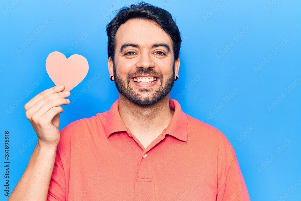 Young hispanic man holding heart shape paper looking positive and happy standing and smiling with a confident smile showing teeth Young hispanic man holding heart shape paper looking positive and happy standing and smiling with a confident smile showing teeth