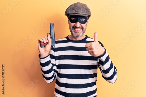 Middle age handsome burglar man wearing cap and mask holding gun over yellow background smiling happy and positive, thumb up doing excellent and approval sign © Krakenimages.com
