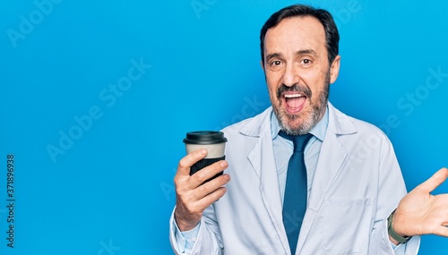 Middle age handsome doctor man wearing coat drinking cup of takeaway coffee celebrating achievement with happy smile and winner expression with raised hand © Krakenimages.com