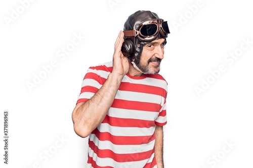 Middle age aviator man wearing vintage helmet and glasses over isolated white background smiling with hand over ear listening and hearing to rumor or gossip. Deafness concept.