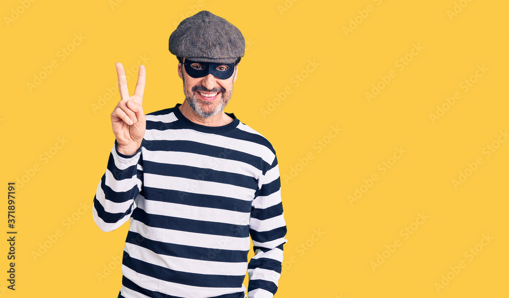 Middle age handsome man wearing burglar mask showing and pointing up with fingers number two while smiling confident and happy. Middle age handsome man wearing burglar mask showing and pointing up with fingers number two while smiling confident and happy.