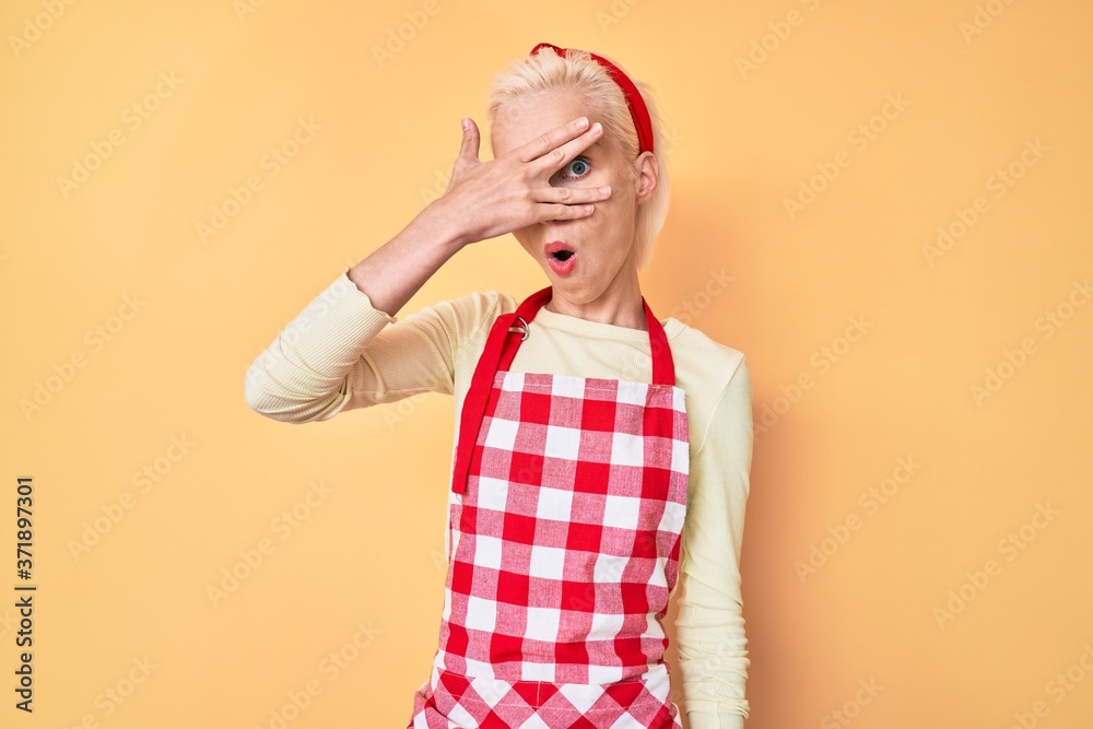 Young blonde woman with tattoo wearing professional baker apron peeking in shock covering face and eyes with hand, looking through fingers afraid Young blonde woman with tattoo wearing professional baker apron peeking in shock covering face and eyes with hand, looking through fingers afraid