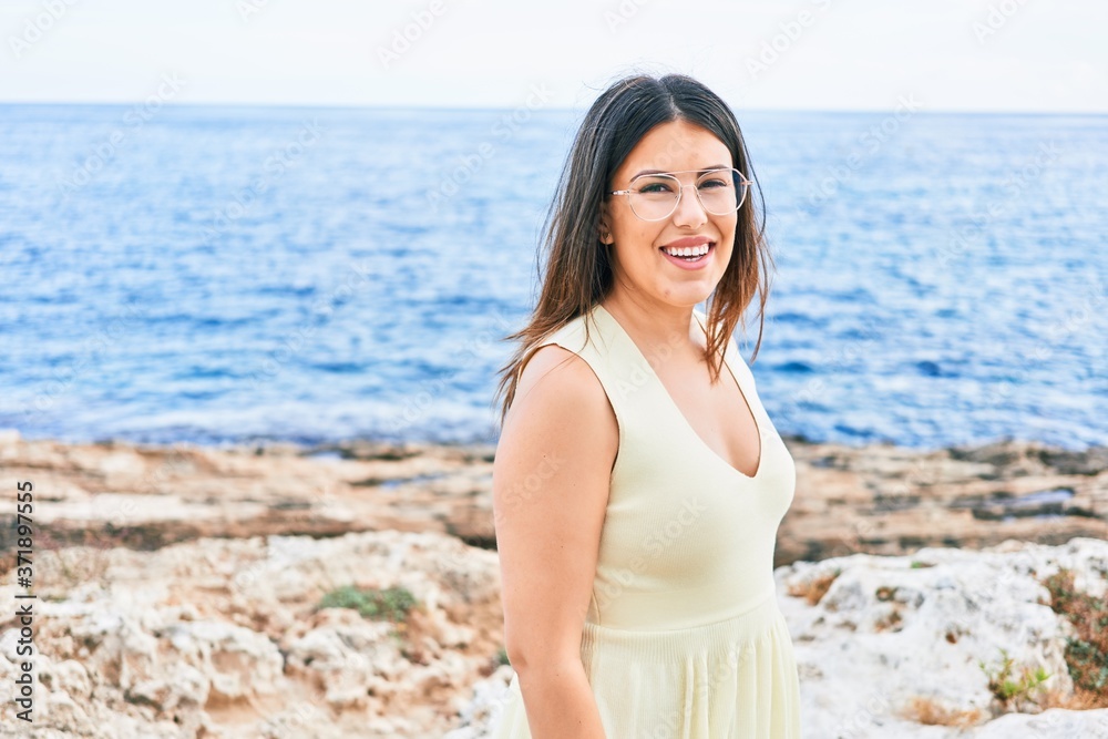 Young beautiful woman smiling happy. Standing with smile on face looking at the camera at the beach Young beautiful woman smiling happy. Standing with smile on face looking at the camera at the beach