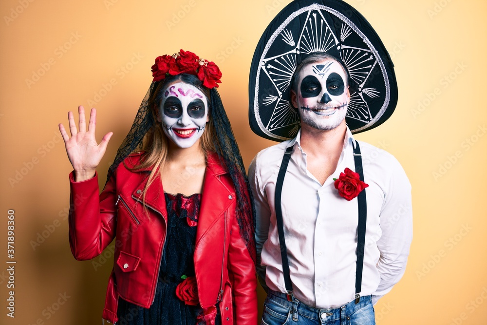 Couple wearing day of the dead costume over yellow showing and pointing up with fingers number five while smiling confident and happy.