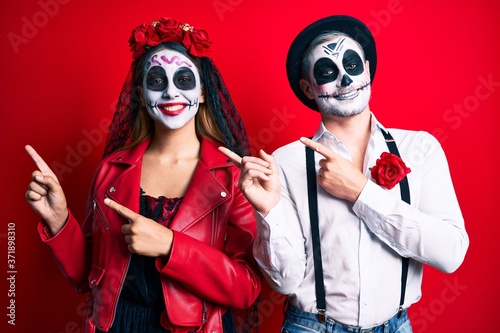 Couple wearing day of the dead costume over red smiling and looking at the camera pointing with two hands and fingers to the side.