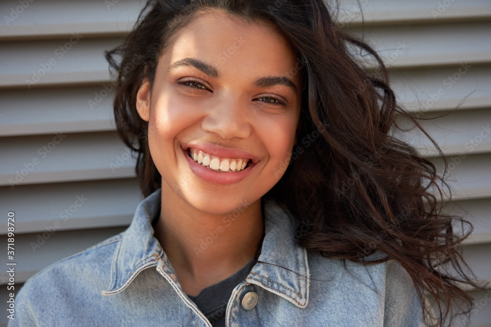 Happy stylish young mixed race gen z girl wearing denim jacket standing outdoor looking at camera, portrait. Pretty millennial African American ethnic woman with dental smile closeup face headshot. - obrazy, fototapety, plakaty 