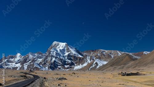A long lonely road with curves on the tibetan plateau with snow clad mountains and peaks and clear blue sky in front and tibetan prayer flags on one side