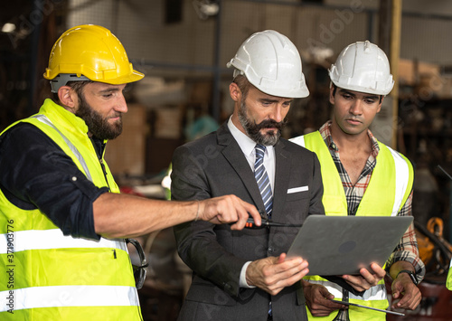 Factory manager in suit using laptop and talking with industrial engineer ,technician man. They wear helmet or hardhat with safety jackets inside industry manufacturing Facility/Discussion  working