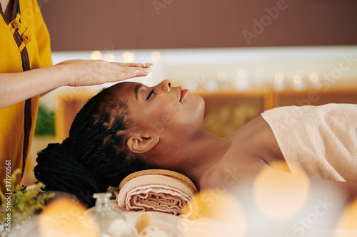 Therapist holding hands over head of pretty young Black woman to transfer energy during healing reiki session photo